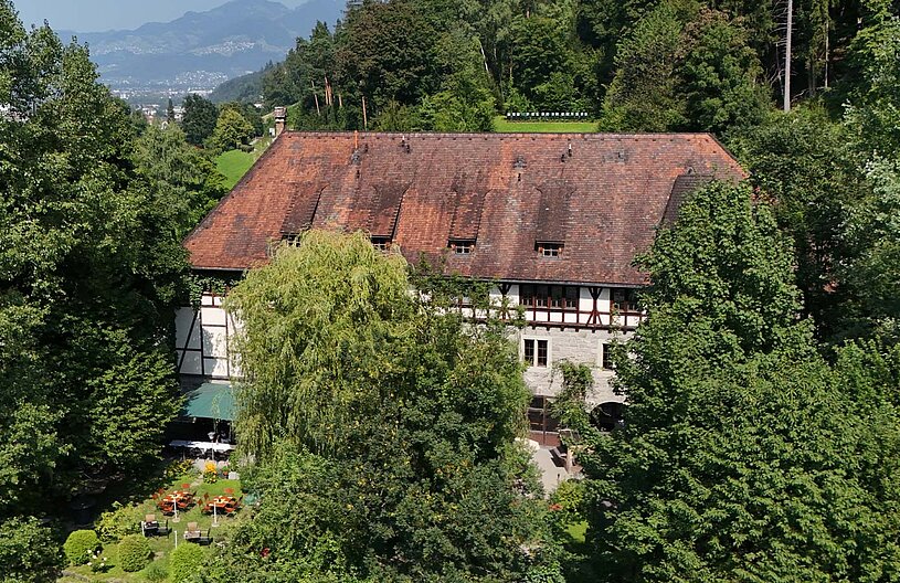 Ein großes, traditionelles Gebäude mit einem schrägen, roten Dach ist von dichtem, grünem Laub umgeben. Vor dem Gebäude sind Sitzgelegenheiten im Freien und ein gepflegter Gartenbereich sichtbar. Im Hintergrund erheben sich sanfte Hügel und eine Berglandschaft.
