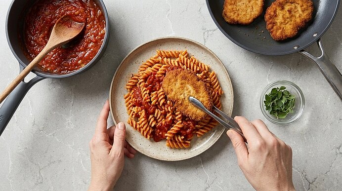 Hände legen ein paniertes Schnitzel mit Zange auf einen Teller Fusilli in Tomatensauce; daneben Pfannen mit Tomatensauce und weiteren panierten Schnitzeln.