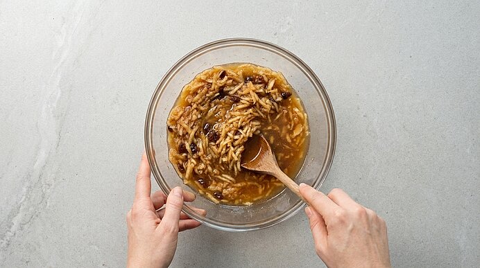 Zwei Hände rühren mit einem Holzlöffel in einer Glasschüssel eine Apfelmischung mit geriebenen Äpfeln, Rosinen und Flüssigkeit, vermutlich als Füllung für Gebäck.