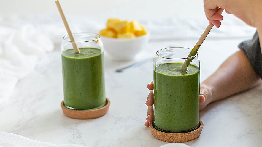 Zwei gläserne Smoothies mit grünlicher Farbe stehen auf einem weißen Tisch. Eine Hand hält ein Glas, während eine andere Hand einen Löffel in den Smoothie taucht. Im Hintergrund befindet sich eine Schüssel mit gelben Fruchtstücken.