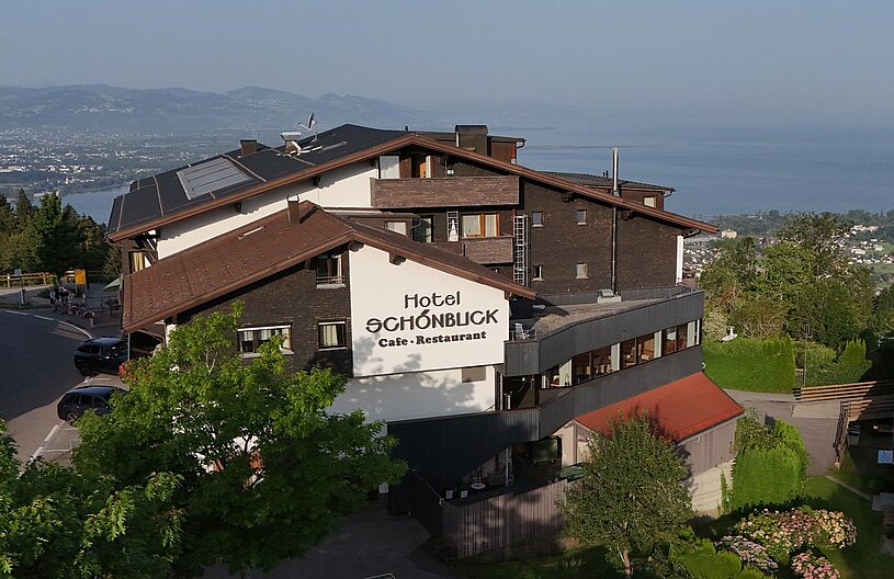 Das Bild zeigt das Hotel Schönblick, das über ein Café und Restaurant verfügt. Es befindet sich auf einem Hang mit Blick auf einen See und die umliegenden Berge. Die Architektur ist modern und umgeben von Bäumen und Grünflächen.