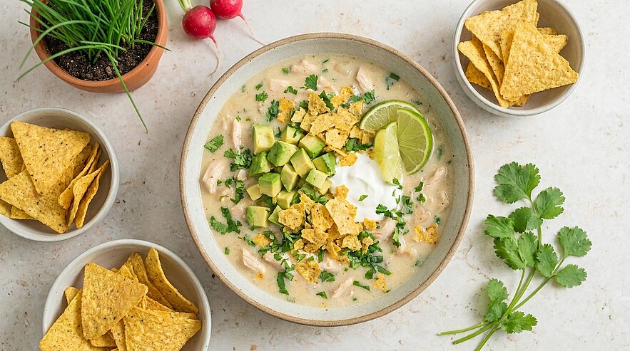 Schüssel cremige weiße Chili mit Hähnchen, gewürfelter Avocado, Limettenscheibe, Sauerrahm, gehacktem Koriander und zerbröselten Tortilla‑Chips; daneben kleine Schälchen mit Nacho‑Chips.