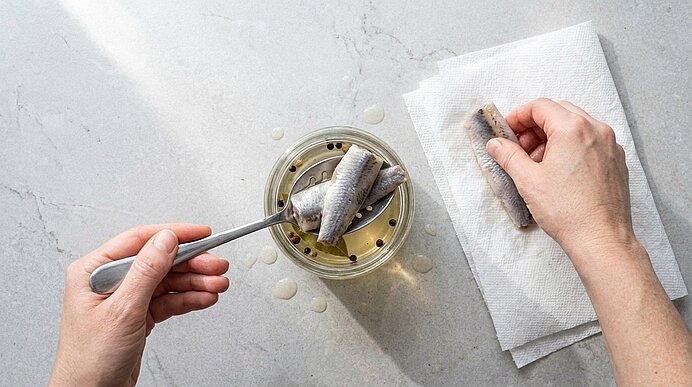 Zwei Hände heben Bismarckhering mit einem Löffel aus einem Glas mit Lake und legen ein Filet auf Küchenpapier.