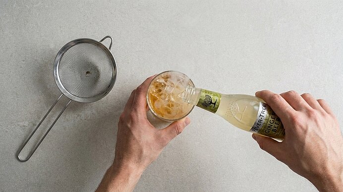 Zwei Hände gießen aus einer Glasflasche ein helles, kohlensäurehaltiges Getränk über Eis in ein Trinkglas.