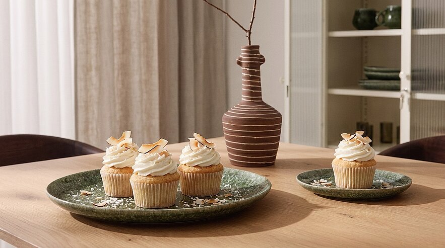Drei Cupcakes mit Sahnehaube und Kokosraspeln auf einem grünen Teller, ein zusätzlicher Cupcake auf einem kleineren Teller daneben. Im Hintergrund steht eine braune Vase. Die Umgebung ist hell und einladend.