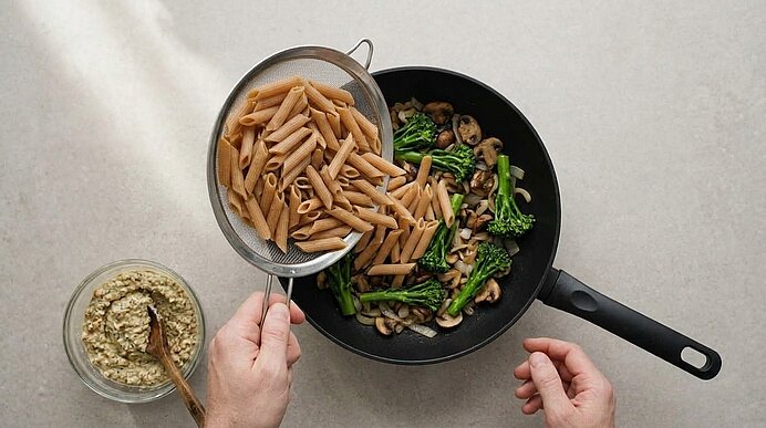 Zwei Hände geben Vollkornpenne aus einem Sieb in eine Pfanne mit Brokkoli und Pilzen. Daneben steht Walnusspesto.