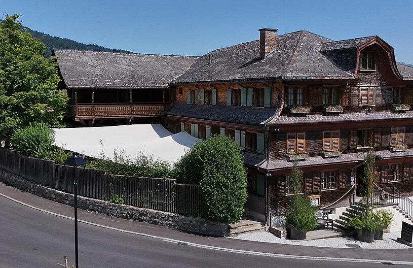 Ein traditionelles Holzgebäude mit mehreren Etagen, großen Fenstern und einem steilen Dach. Umgeben von Bäumen und Pflanzen, steht eine weiße Markise im Vordergrund. Die Straße verläuft halbkreisförmig um das Haus. Im Hintergrund sind sanfte Hügel zu sehen.