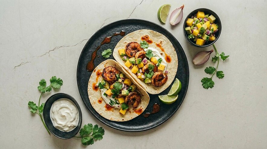 Zwei Garnelen-Tacos auf dunklem Teller, gefüllt mit Mango‑Salsa, Koriander und Limetten, daneben Schälchen mit Sauce und saurer Creme.