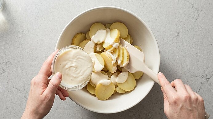 Zwei Hände geben cremiges Dressing aus einem Glas über in Scheiben geschnittene Kartoffeln in einer weißen Schüssel; ein Spatel rührt die Mischung.