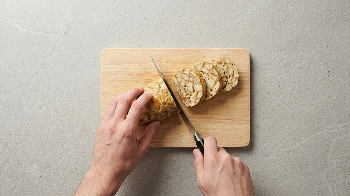 Hände schneiden mit einem Messer eine längliche Brot- oder Teigrolle in Scheiben auf einem Holzbrett.