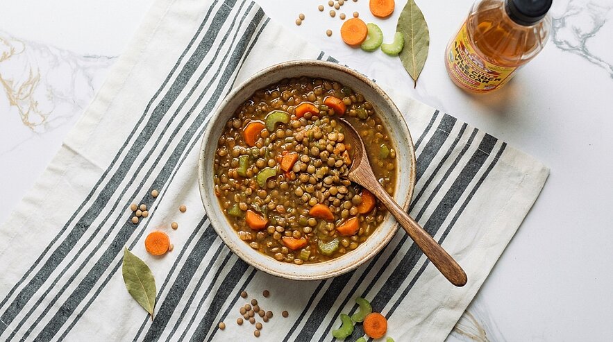 Schale mit herzhafter Linsensuppe, erkennbar Linsen, Karottenscheiben und Selleriestücke, darin ein Holzlöffel; steht auf gestreiftem Tuch.