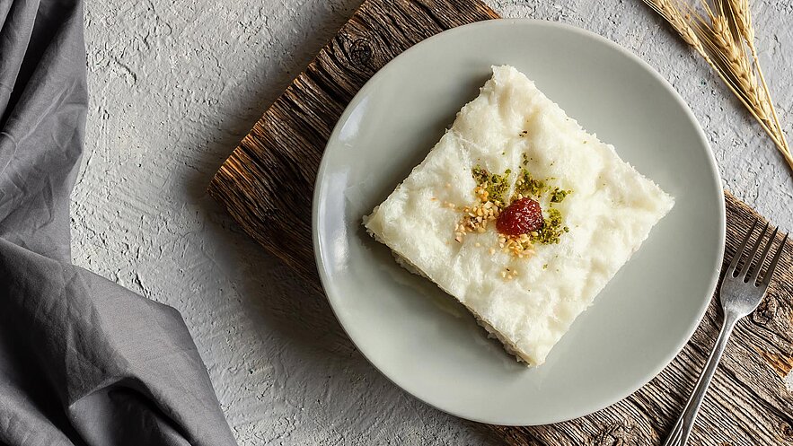 Ein Stück Dessert auf einem grauen Teller, garniert mit gehackten Pistazien und einer roten Frucht. Der Teller steht auf einem rustikalen Holzbrett. Das Dessert hat eine weiche, cremige Textur und ist ansprechend präsentiert.