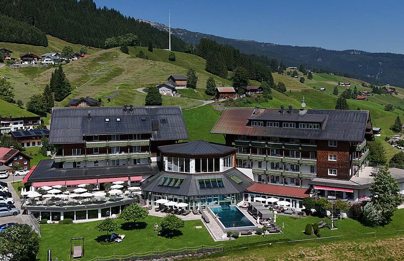 Ein modernes Hotel mit einem Pool liegt in einer malerischen Berglandschaft. Umgeben von grünen Wiesen und Bäumen, sind im Vordergrund Sonnenschirme und Liegen zu sehen. Im Hintergrund erheben sich sanfte Hügel und Wälder.