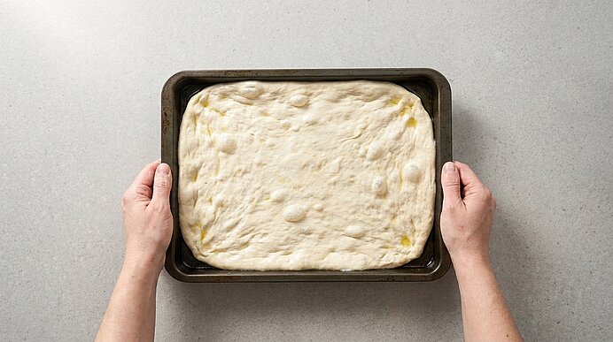Zwei Hände halten ein mattschwarzes Backblech mit ruhendem Pizzateig auf einer hellgrauen Steinarbeitsplatte.