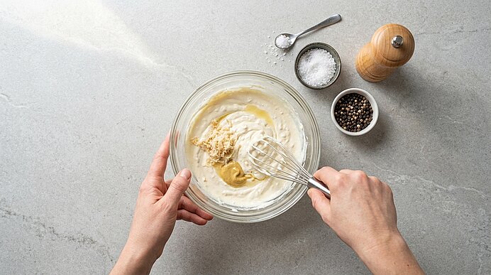 Zwei Hände verrühren Crème fraîche, Frischkäse, Meerrettich und Senf mit einem Schneebesen in einer Glasschüssel.