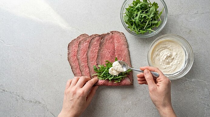 Zwei Hände geben Meerrettichcreme per Löffel auf Rucola auf eine Roastbeefscheibe. Daneben stehen zwei Glasschüsseln.