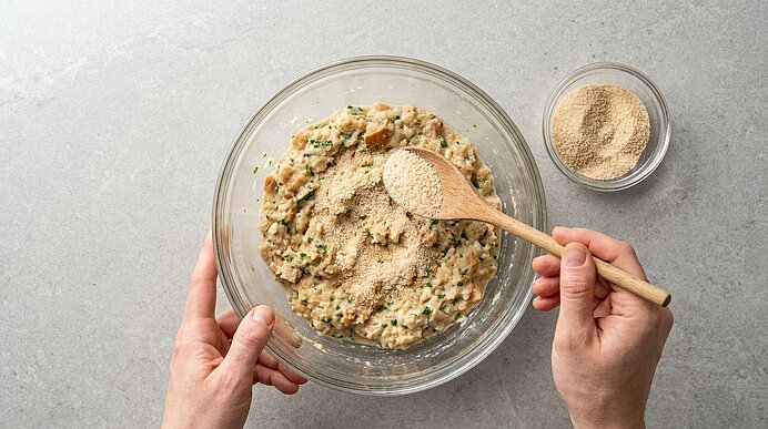 Hände geben mit einem Holzlöffel Semmelbrösel in eine große Glasschüssel mit gewürzter Mischmasse und Kräutern, offenbar zur Zubereitung von Bratlingen oder Frikadellen.
