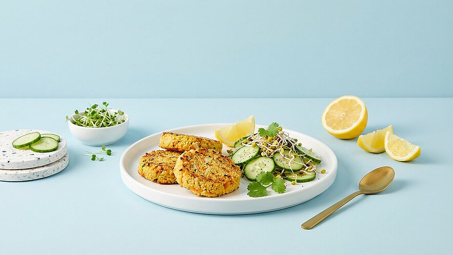 Auf einem Teller liegen zwei goldbraune Bratlinge neben einem frischen Salat aus Gurkenscheiben, Sprossen und Koriander. Das Gericht wird von Zitronenscheiben und einem kleinen Schälchen mit Sprossen begleitet. Der Hintergrund ist in sanftem Blau gehalten.
