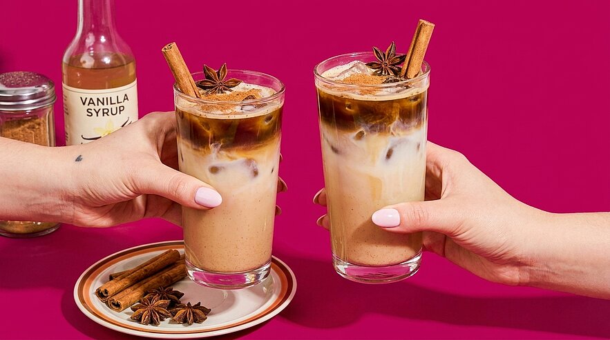 Zwei Hände stoßen mit hohen Gläsern Eiskaffee mit Eis, Zimtstangen und Sternanis an; Vanillesirupflasche und Gewürze im Hintergrund.