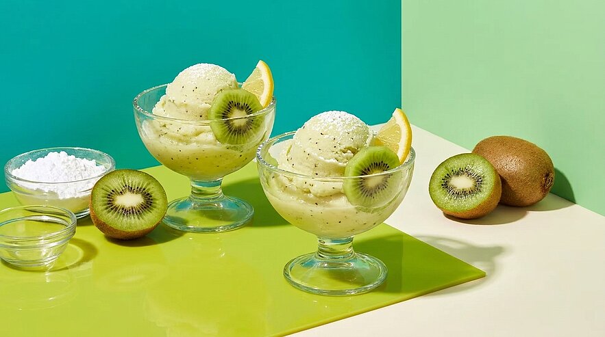 Zwei Glasdessertschalen mit hellgrünem Kiwi-Sorbet, jeweils garniert mit einer Kiwischeibe und Zitronenschnitz; daneben liegen eine halbierte und zwei ganze Kiwis.