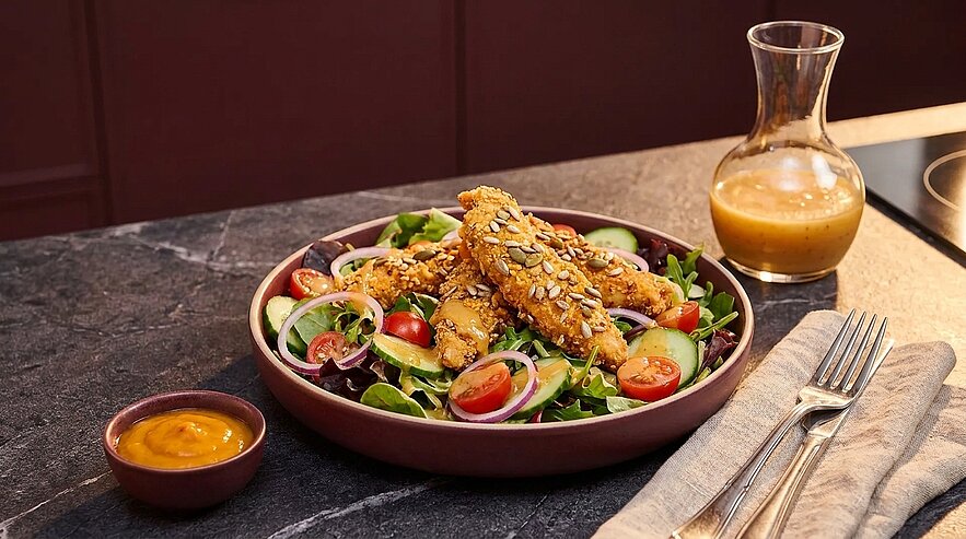 Teller mit gemischtem Salat (Blattsalat, Kirschtomaten, rote Zwiebeln, Gurkenscheiben) belegt mit knusprigen, körnigen Panierstreifen; daneben ein kleines Schälchen Sauce und ein Kännchen Dressing.