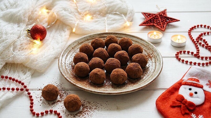 Ein Teller mit mehreren kakaoüberzogenen Schokoladen-Trüffeln auf weißem Tisch, umgeben von Weihnachtslichtern, Teelichtern, einem roten Stern und roter Perlenkette.