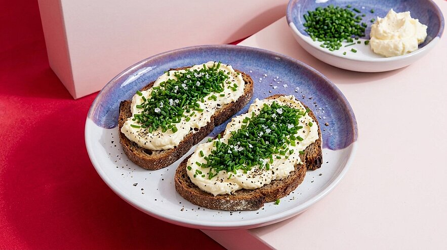 Zwei Scheiben dunkles Mehrkornbrot mit cremigem Frischkäseaufstrich und reichlich gehacktem Schnittlauch auf einem Teller; im Hintergrund ein Schälchen mit extra Schnittlauch und Aufstrich.