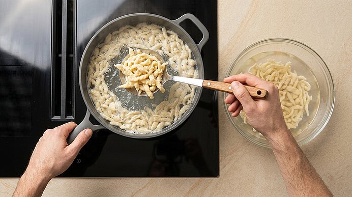 Zwei Hände schöpfen frisch gekochte Spätzle mit einem Löffel aus einer Pfanne auf dem Herd in eine Glasschüssel.