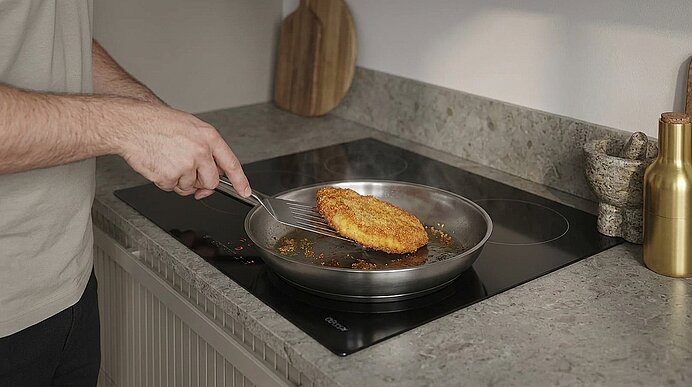 Person wendet ein paniertes Schnitzel mit Pfannenwender in einer Bratpfanne auf einem Glaskeramikkochfeld.