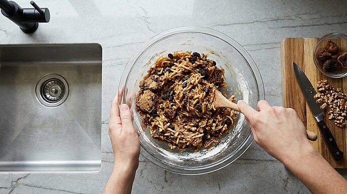 Zwei Hände rühren mit einem Holzlöffel in einer großen Glasschüssel einen Teig mit Nüssen und Trockenfrüchten auf einer Küchenarbeitsplatte.