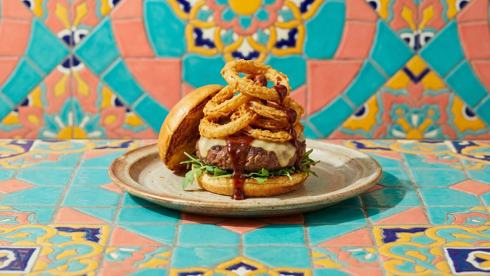 Ein saftiger Burger mit geschmolzenem Käse, belegt mit Röstzwiebeln und einer Barbecue-Sauce, ruht auf einem Teller. Im Hintergrund ist eine bunte, geflieste Wand zu sehen. Der Burger ist von frischem Salat umgeben.