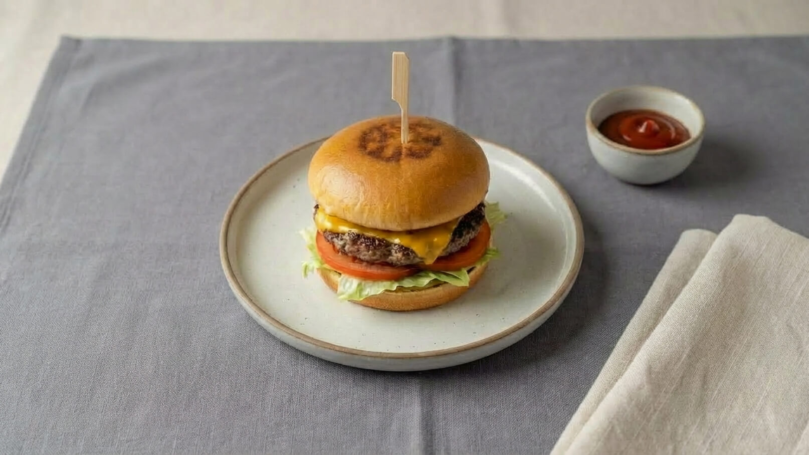 Ein frisch zubereiteter Hamburger auf einem Teller. Der Burger besteht aus einem weichen Brötchen, einer saftigen Fleischpatty, geschmolzenem Käse, frischen Tomatenscheiben und knackigem Salat. Neben dem Teller befindet sich eine kleine Schüssel mit Ketchup.