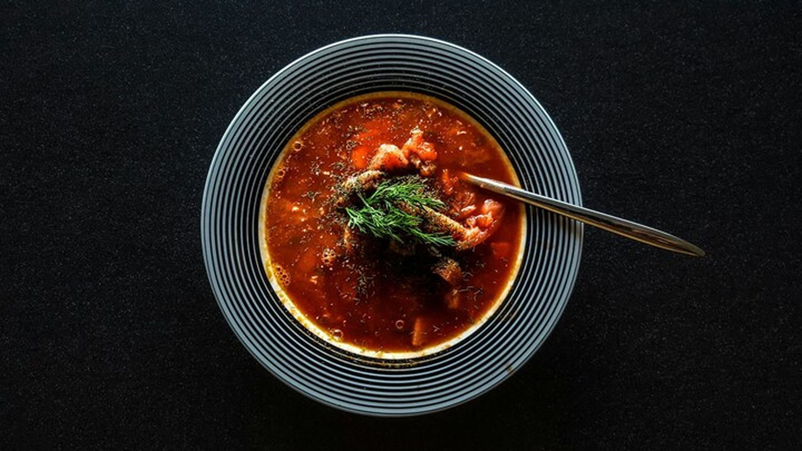 Schüssel mit dampfender Tomatensuppe, garniert mit Fleischstücken und frischen Kräutern. Der Rand der Schüssel ist gestreift und der Löffel liegt daneben. Der Hintergrund ist dunkel und hebt die Farben der Suppe hervor.