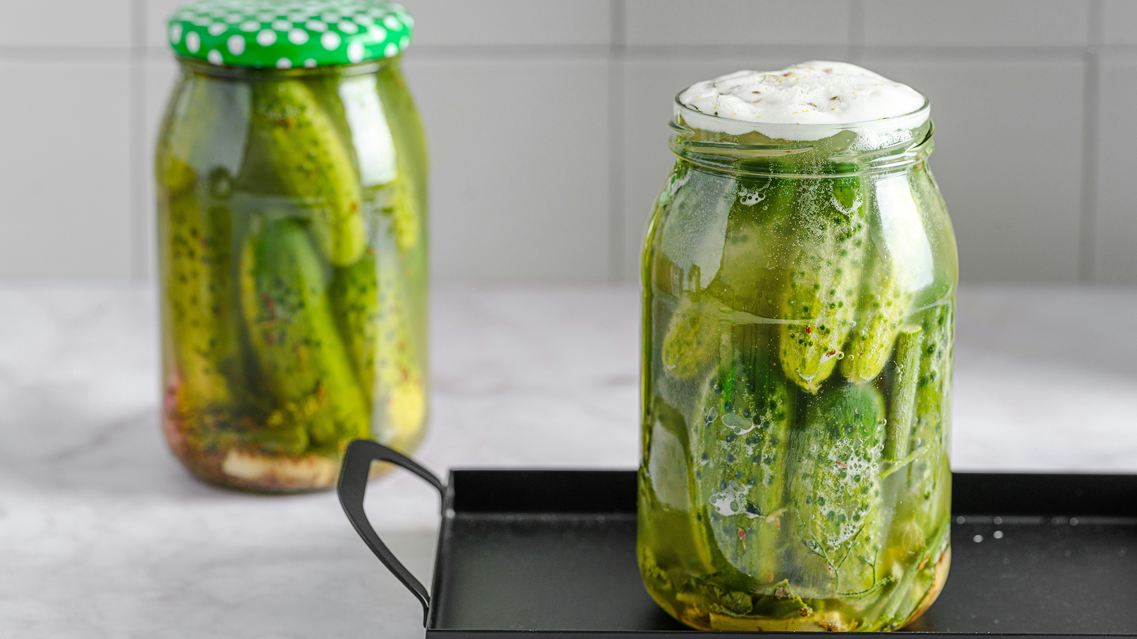 Zwei transparente Gläser mit eingelegten Gurken stehen auf einer Tischoberfläche. Das vordere Glas ist voll mit grünen Gurken und einer schaumigen Flüssigkeit, während das hintere Glas einen grünen Deckel hat und ähnliche Gurken enthält.