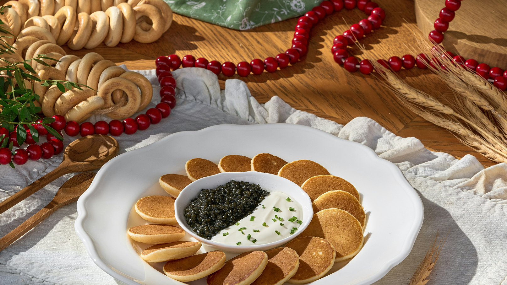 Eine weiße Schale auf einem Tisch, gefüllt mit einem Dip aus saurer Creme und schwarzem Kaviar in der Mitte. Rundherum sind dünne, goldbraune Pfannkuchen angeordnet. Im Hintergrund sind dekorative Elemente wie Gebäck und eine rote Perlenkette zu sehen.