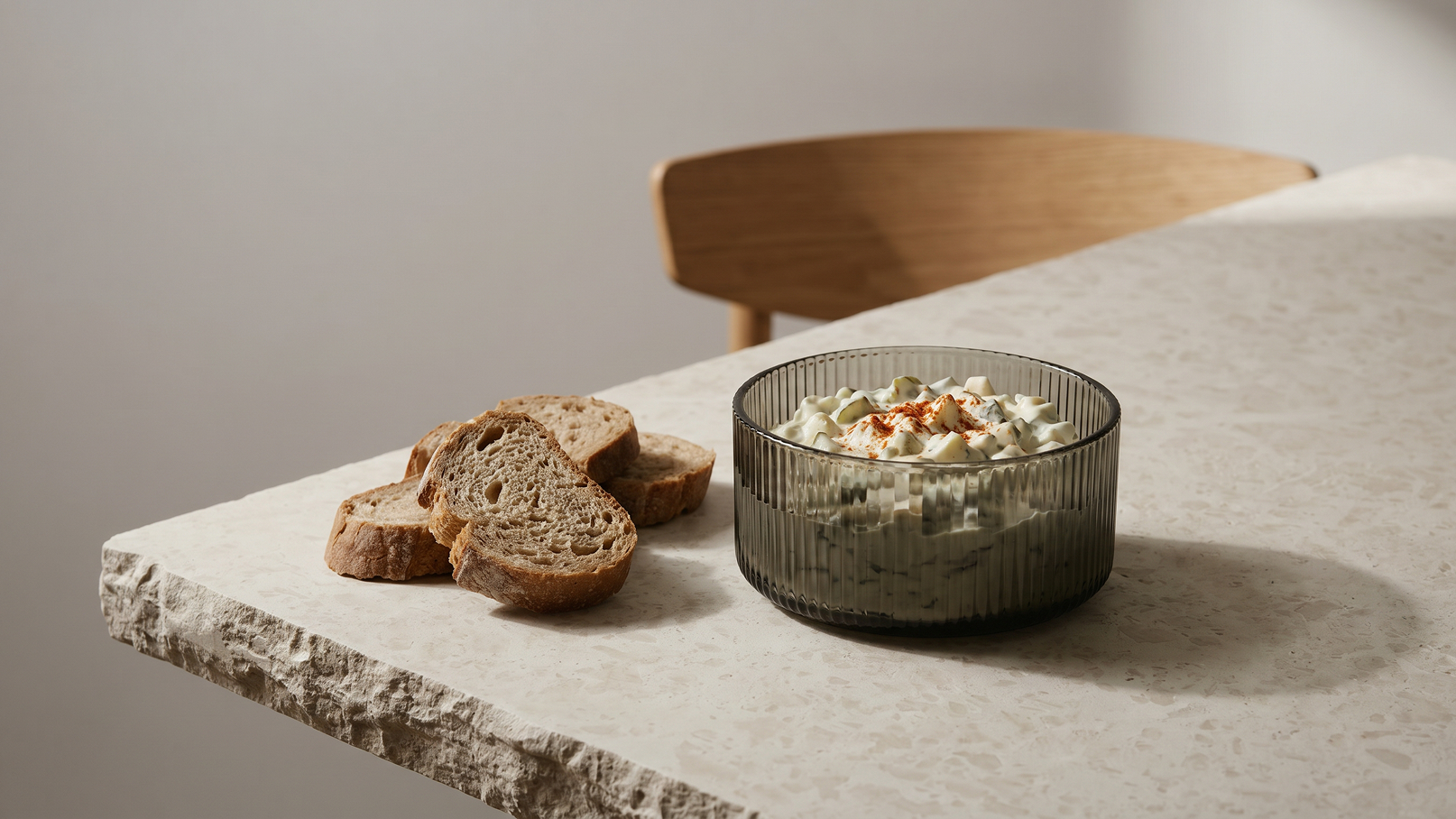 Auf einem Tisch liegt ein Glas mit einer cremigen Speise, garniert mit Gewürzen. Daneben befinden sich mehrere Scheiben dunkles Brot. Der Tisch hat eine helle, steinartige Oberfläche, und im Hintergrund ist ein Stuhl aus Holz zu sehen.