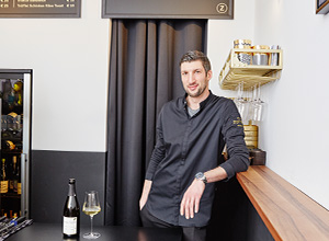 Ein lächelnder Barkeeper steht an einer Theke und lehnt sich leicht gegen sie. Vor ihm steht ein Glas und eine Flasche Wein. Im Hintergrund sind Regale mit Getränken zu sehen. Die Atmosphäre wirkt modern und einladend.
