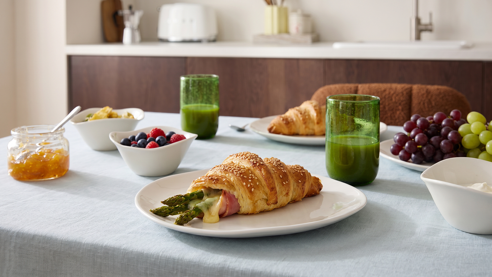 Ein gedeckter Tisch mit einem Croissant, gefüllt mit Schinken und Spargel, auf einem weißen Teller. Daneben stehen Schalen mit frischen Beeren, Trauben und anderen Früchten sowie Gläser mit grünem Saft. Im Hintergrund ein Glas mit Marmelade.