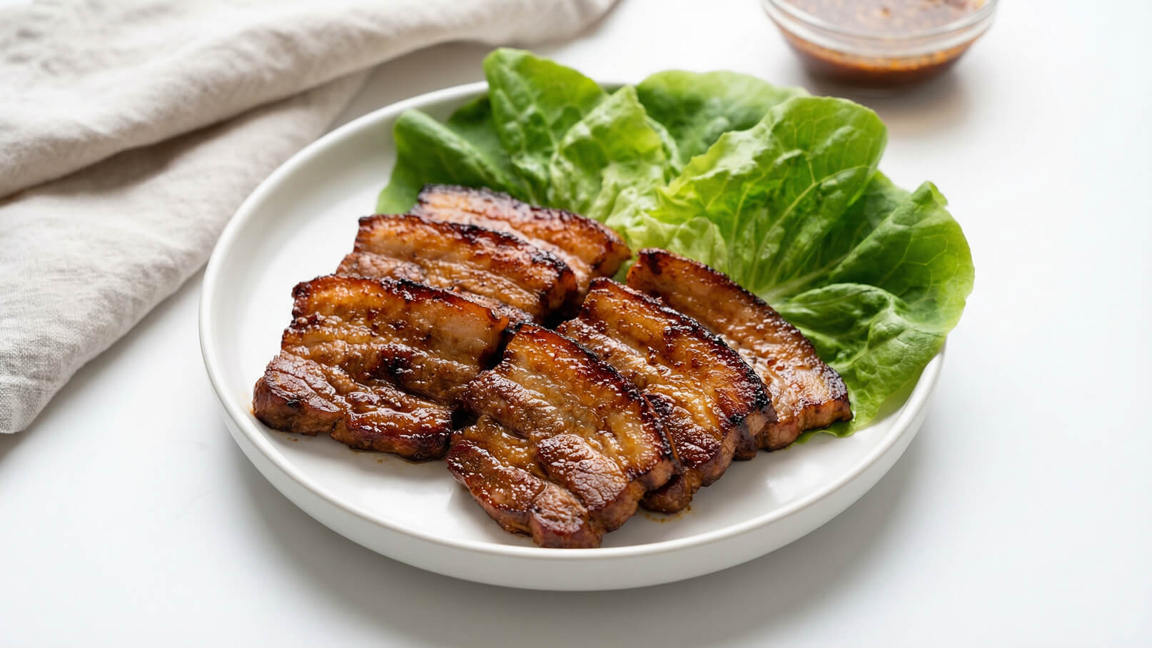Auf dem Bild ist ein Teller mit gegrilltem Fleisch zu sehen, das in gleichmäßige Scheiben geschnitten ist. Neben dem Fleisch liegen frische Salatblätter. Im Hintergrund befindet sich eine kleine Schale mit einer Sauce.