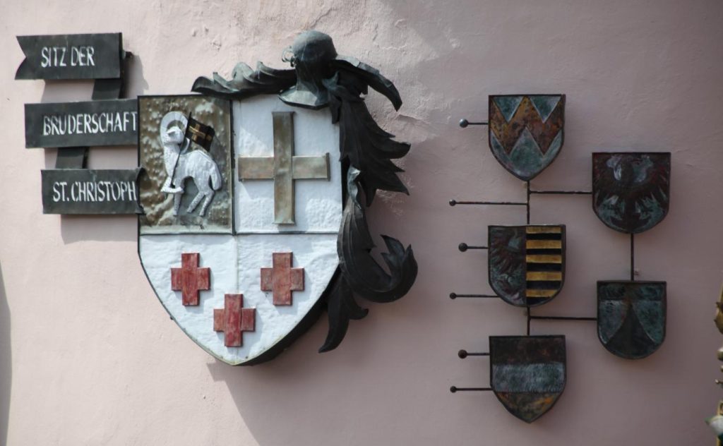 Das Bild zeigt ein Wappen der Bruderschaft St. Christoph mit einem traditionellen Design. Es enthält ein Kreuz, ein Pferd und rote Kreuzsymbole. Daneben sind mehrere kleinere Wappen in unterschiedlichem Design angeordnet, die zu der zentralen Darstellung gehören.