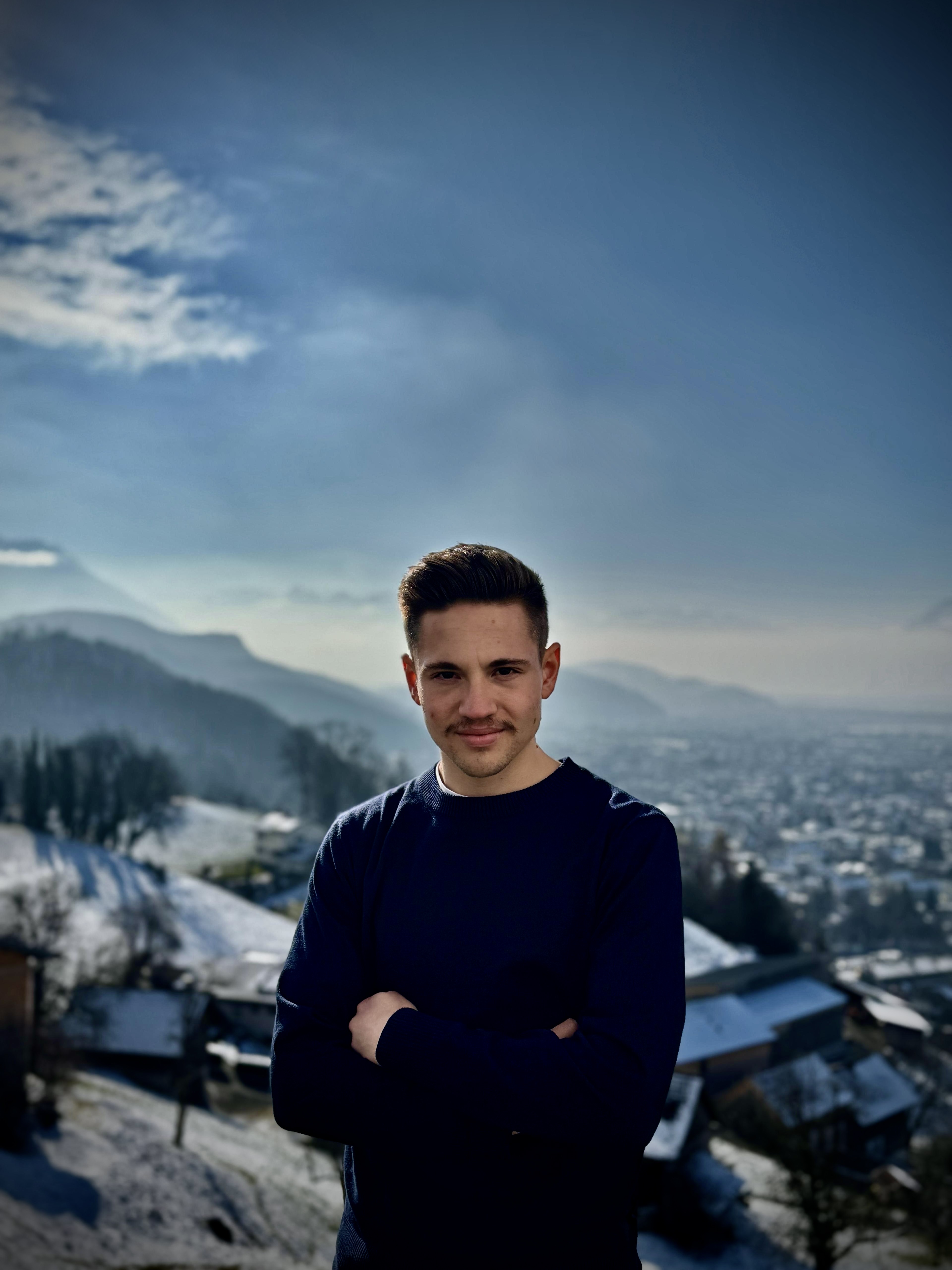 Ein junger Mann steht mit verschränkten Armen vor einer winterlichen Landschaft. Im Hintergrund sind schneebedeckte Hügel und eine weite Aussicht auf eine Stadt sichtbar. Der Himmel ist klar und blau, was eine ruhige und friedliche Stimmung vermittelt.
