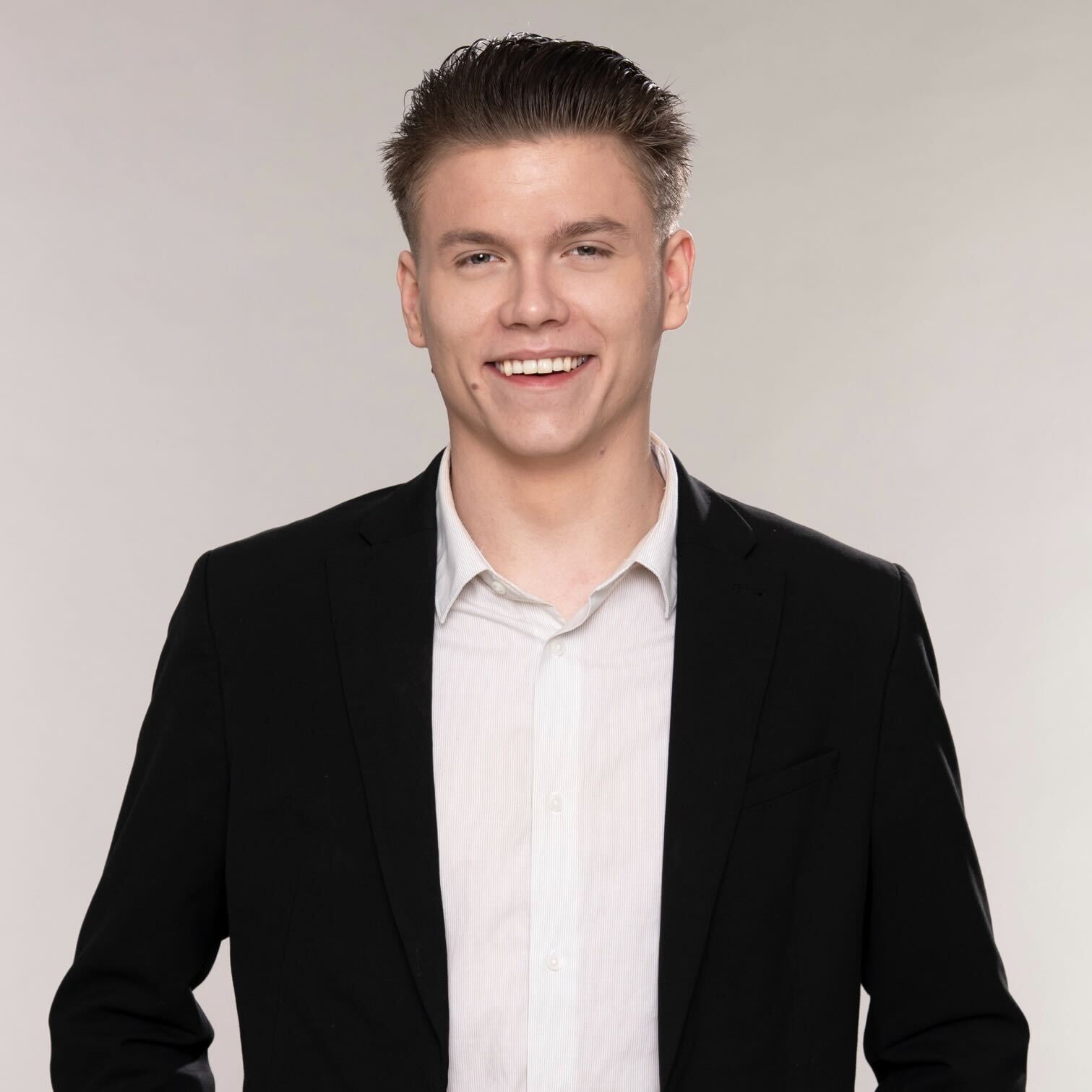 Ein junger Mann mit lässigem Lächeln trägt einen dunklen Anzug und ein weißes Hemd. Er steht vor einem hellen Hintergrund und vermittelt einen freundlichen und professionellen Eindruck.