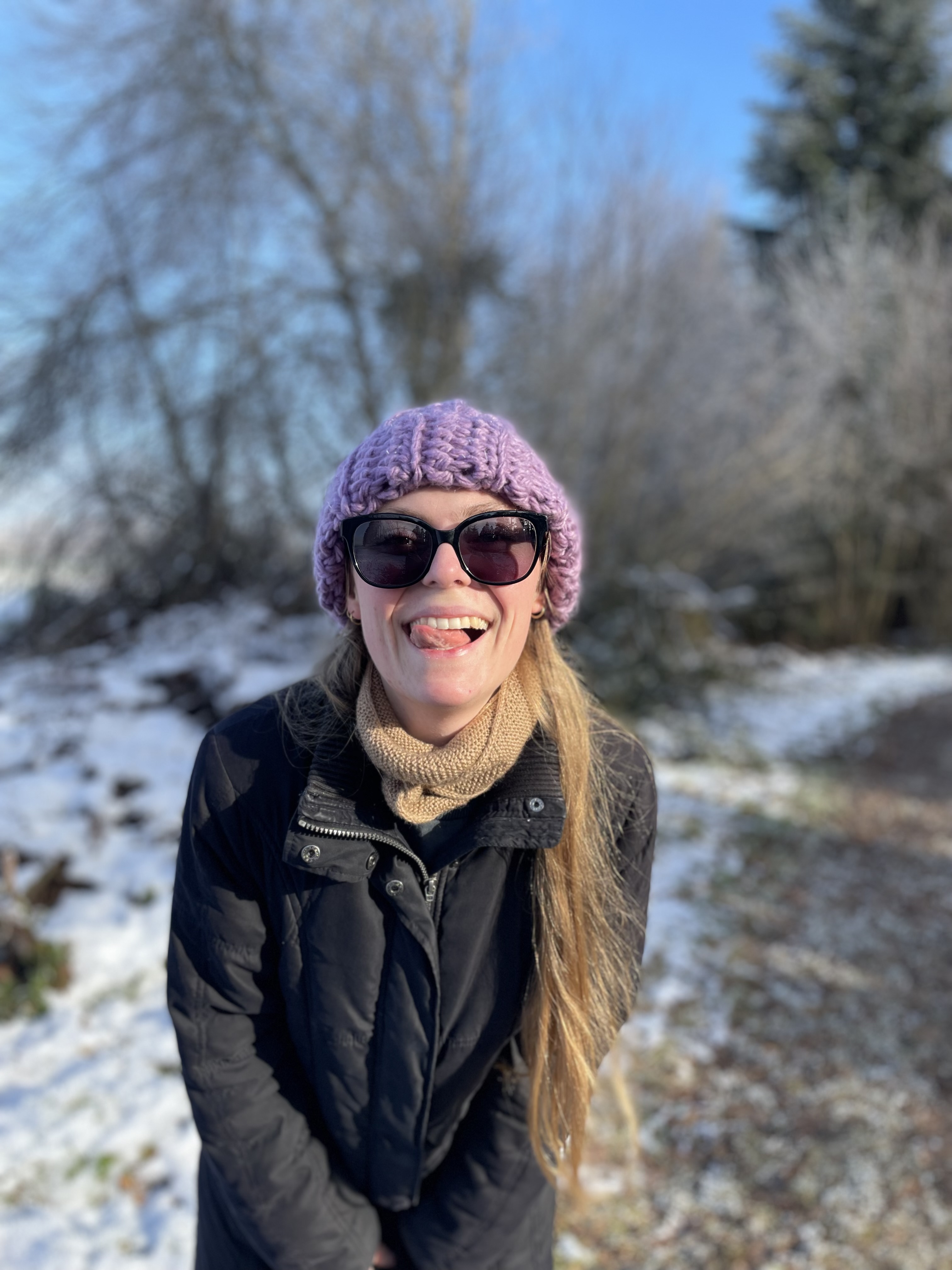 Eine Person mit langen, blonden Haaren trägt eine lila Mütze und eine schwarze Jacke. Sie steht draußen auf einem verschneiten Weg, lächelt und stickt die Zunge heraus. Im Hintergrund sind Bäume und blauer Himmel sichtbar.